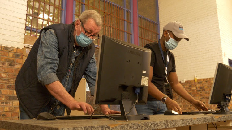 Photo: two middle aged men in vests and surgical masks in front of standing, typing on a computer.